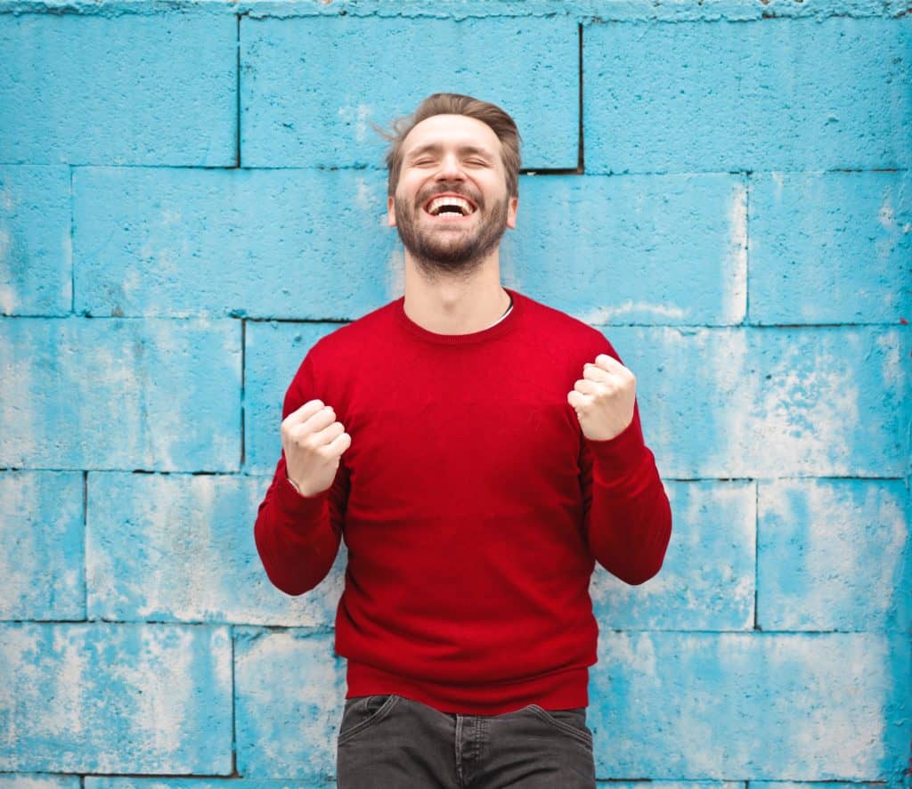 A man in a red sweater is celebrating against a blue wall while showcasing his PPC Management Certification.